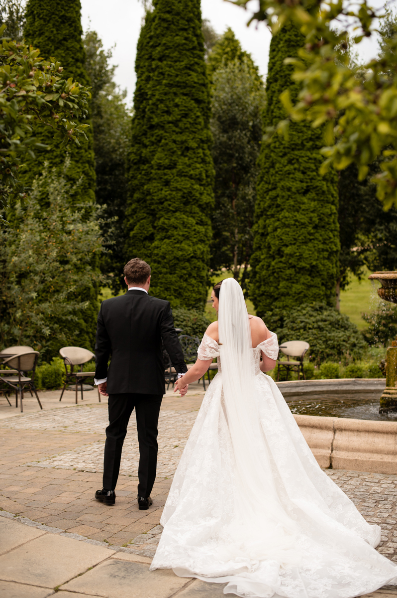 Bride and groom walking hand in hand through the landscaped gardens at Crossbasket Castle after their wedding ceremony.