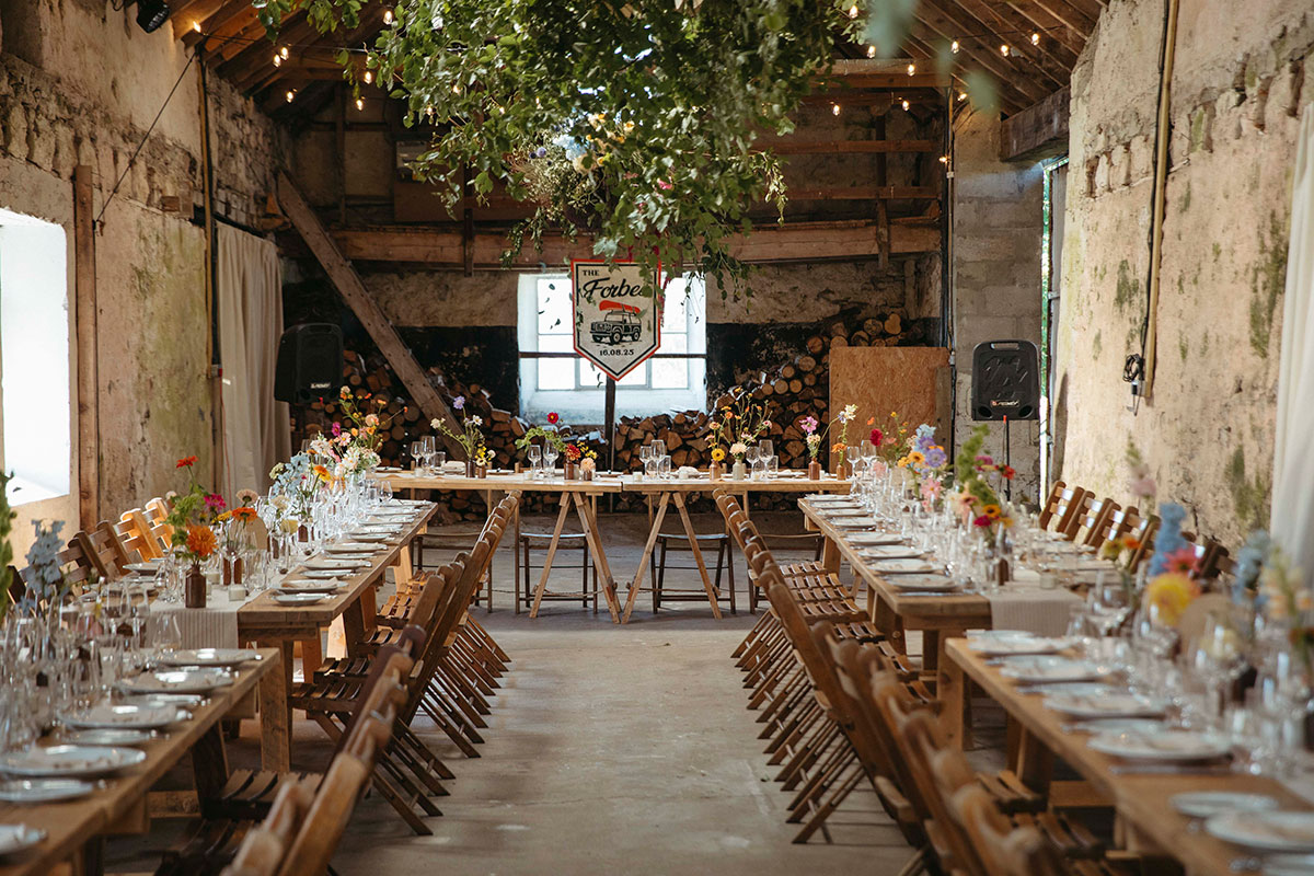 Rustic barn wedding reception set with long wooden tables, colourful wildflowers, hanging greenery and festoon lights.