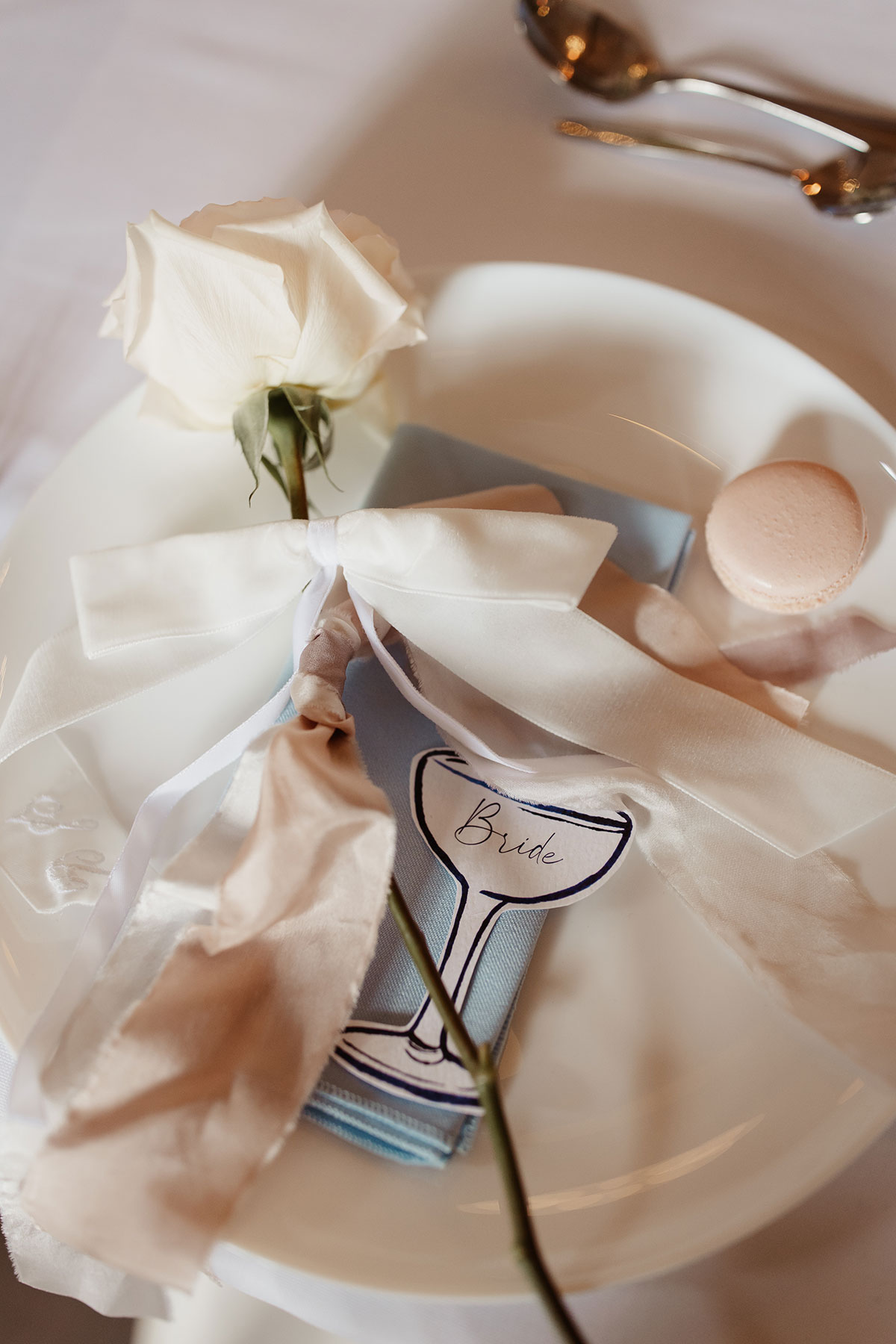 a champagne coupe illustrated place setting with blue napkin, pink macaron and ivory ribbon bow around a single ivory rose stem