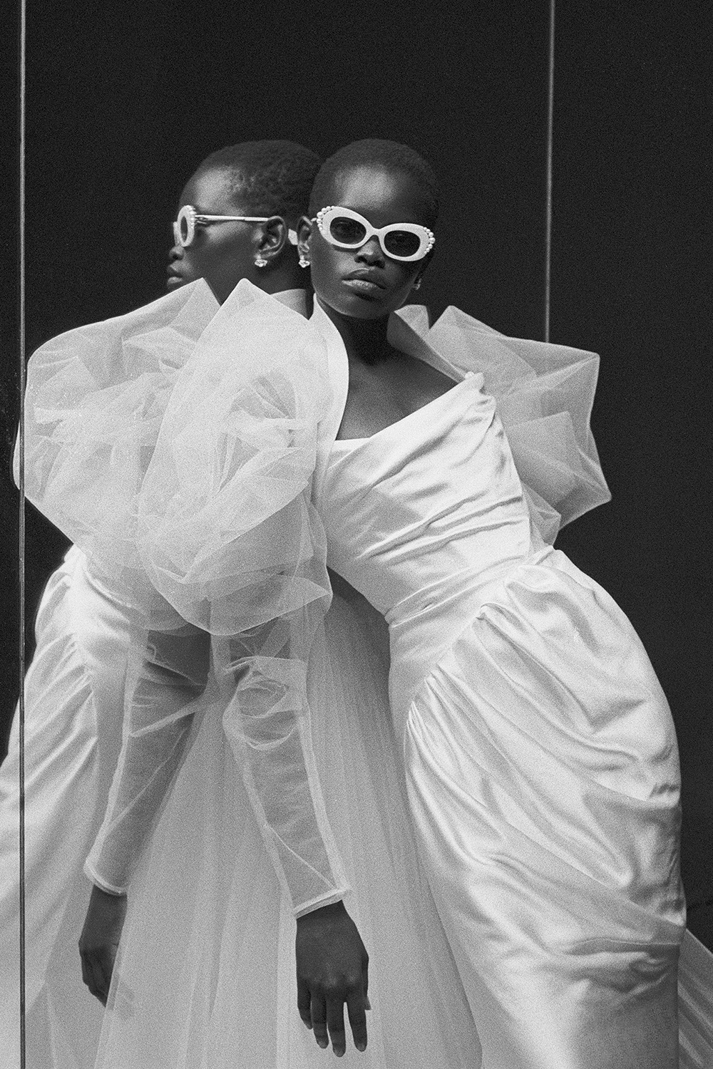 Model in white dress and sunglasses leaning against a mirror