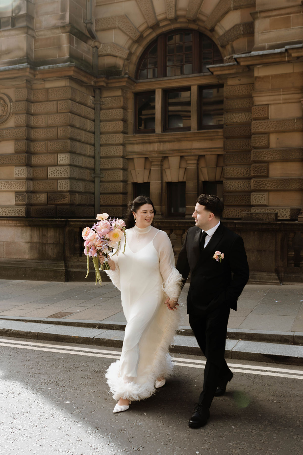 Newlyweds in wedding attire walking hand in hand outside a historic sandstone building in a Scottish city
