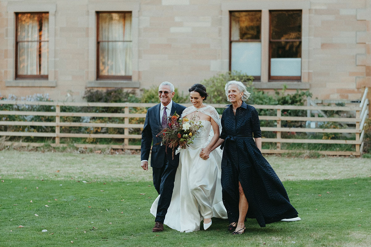 Bride walks across the lawn with her parents ahead of her outdoor wedding ceremony at Cambo Country House in Fife.