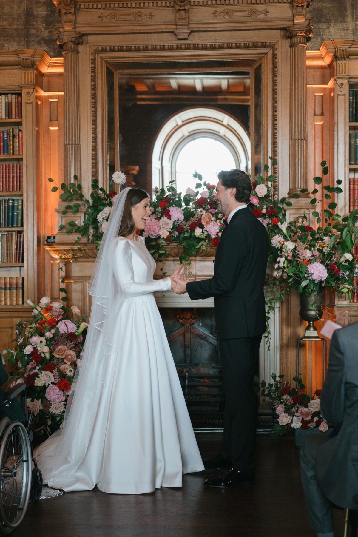 Nicole and Ryan exchange vows during their wedding ceremony in the library at Barnbougle Castle, surrounded by pink-and-red floral arrangements