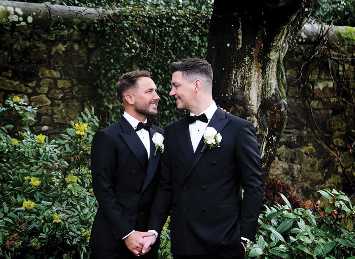 two grooms hold hands and smile at one another. There is a stone wall, a tree trunk and lots of greenery in the background