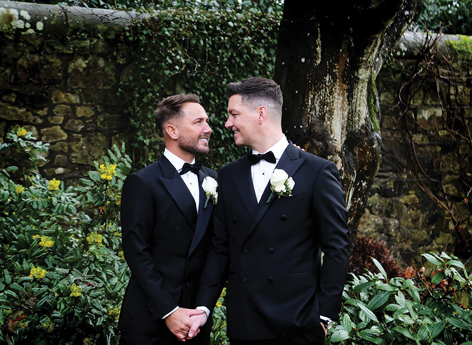 two grooms hold hands and smile at one another. There is a stone wall, a tree trunk and lots of greenery in the background