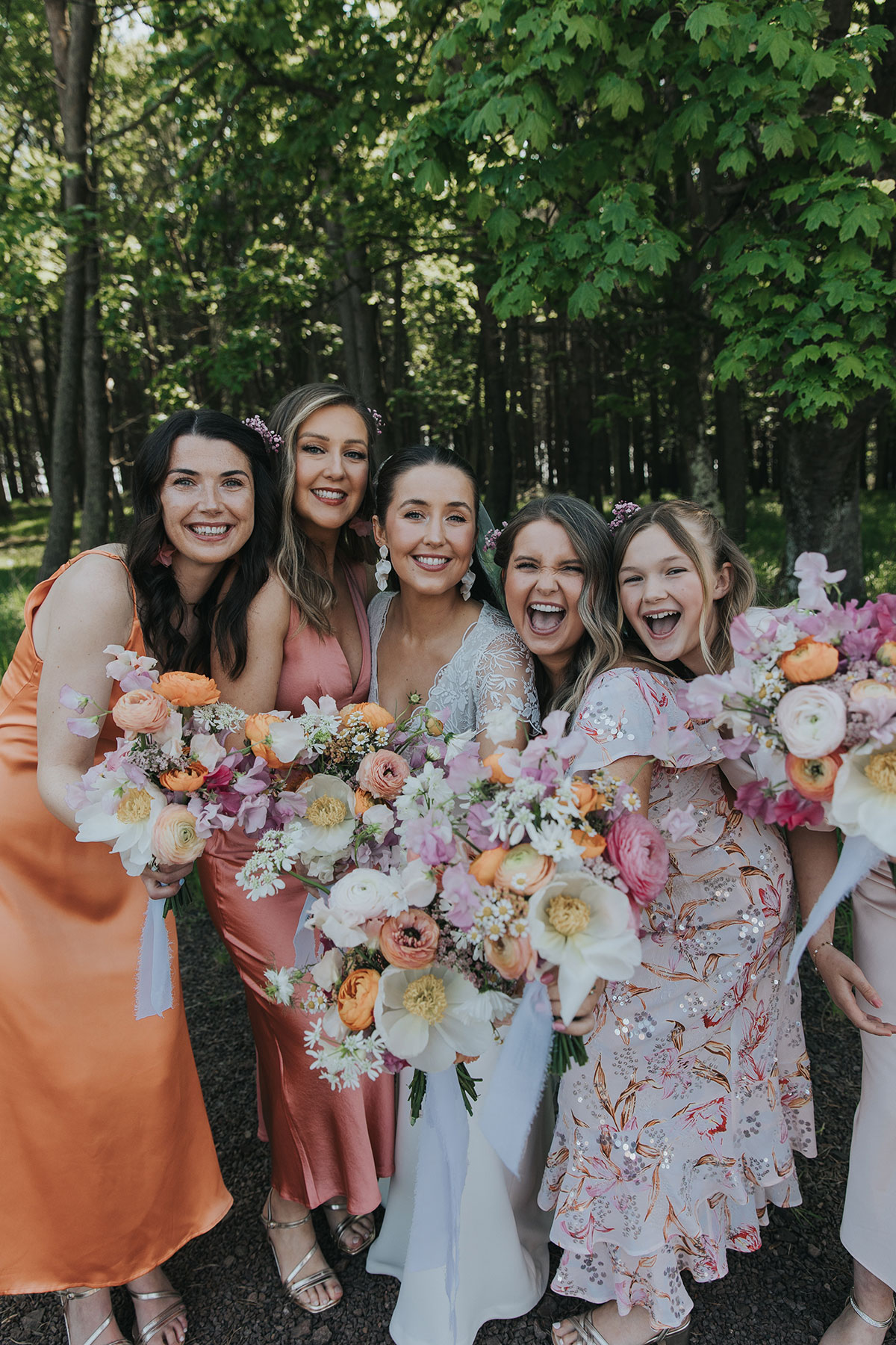 bride and bridesmaids wearing mismatched colourful dresses hold up bouquets towards the camera as they smile