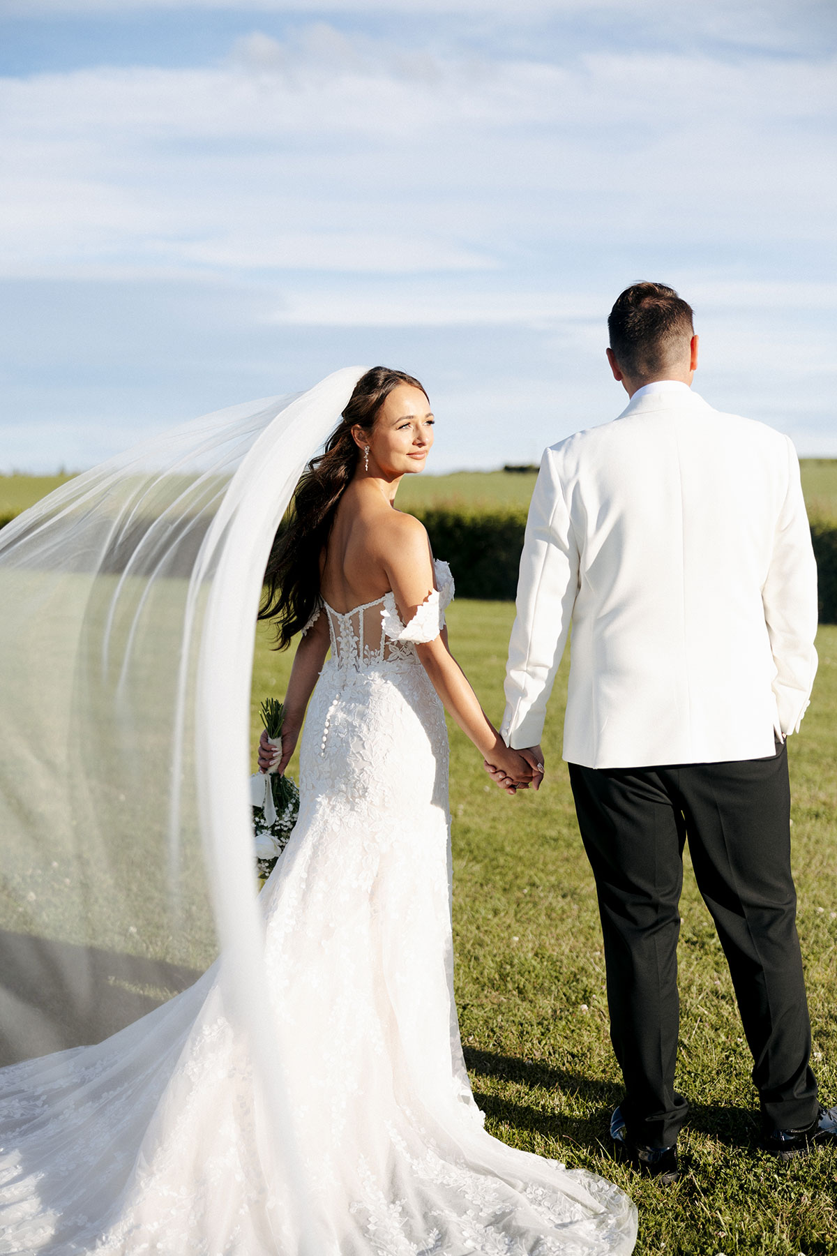 Newlyweds stroll through a sunlit field at Falside Mill, holding hands and smiling against the blue sky backdrop.