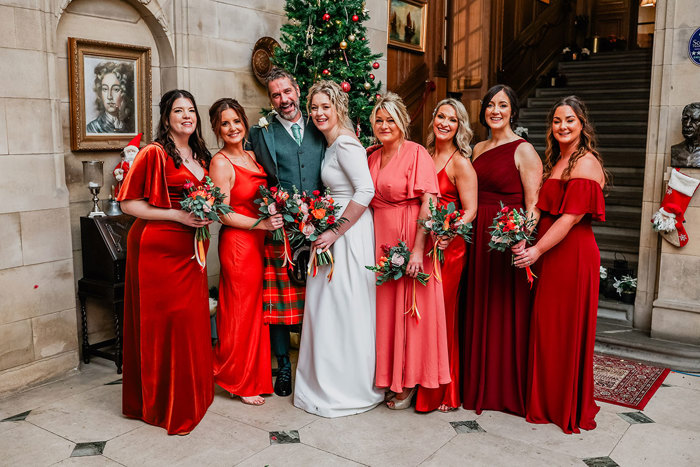 bride in white wedding dress and groom in red and green tartan kilt outfit stand in front of Christmas tree with a bridal party of six women by either sides of them in varying shades of red dresses