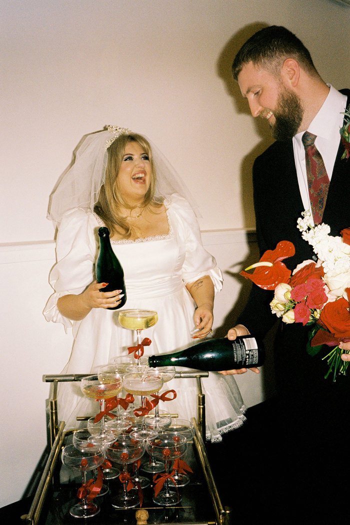 bride and groom laugh as they each pour a bottle of champagne into a tower of coupe glasses, all of which are decorated with red bows and placed on a gold cocktail trolley