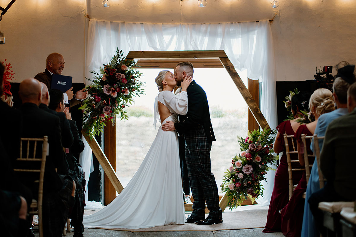 A bride and groom kissing at the end of their ceremony beneath a wooden arch decorated with pink and burgundy flowers.
