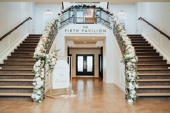 Double staircase at the Firth Pavilion at Seamill Hydro decorated with pink and white flowers on bannisters with gold and white wedding welcome sign 