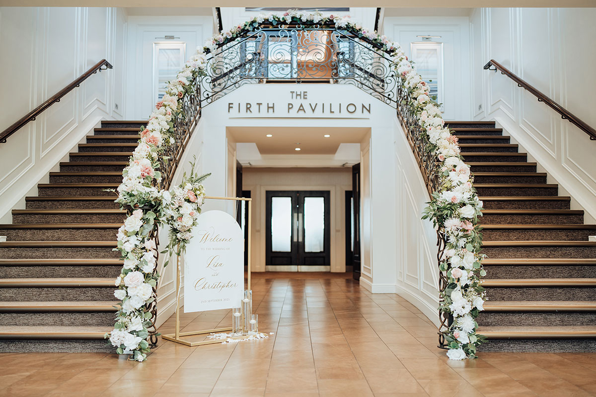 Double staircase at the Firth Pavilion at Seamill Hydro decorated with pink and white flowers on bannisters with gold and white wedding welcome sign