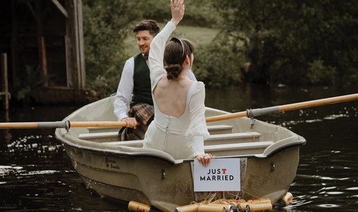 bridal couple sat in a row boat with a just married sign and beer cans hanging off the back of the boat