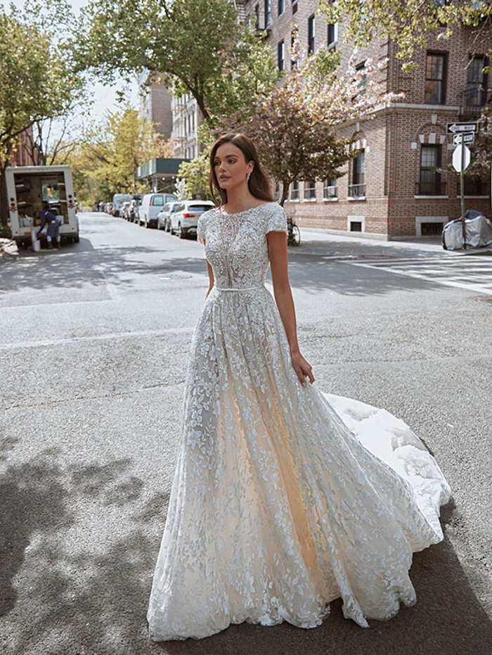 Woman walks across street wearing short-sleeved floor-length wedding dress detailed with floral embellishments 