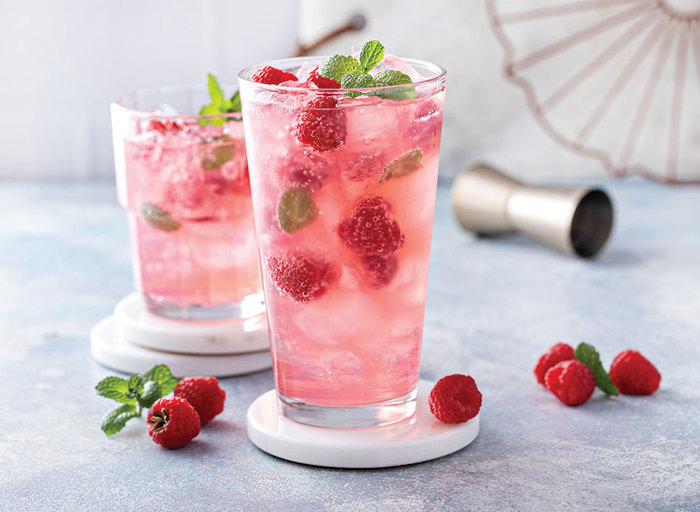 A pink drink in two tall glasses with raspberries inside the glasses and surrounding them 