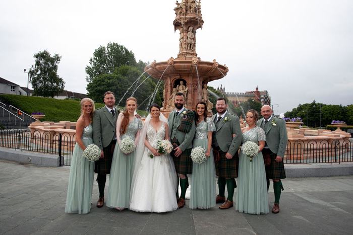 a bridal party stand in front of a fountain with the men in green kilts and the bridesmaids in sage dresses