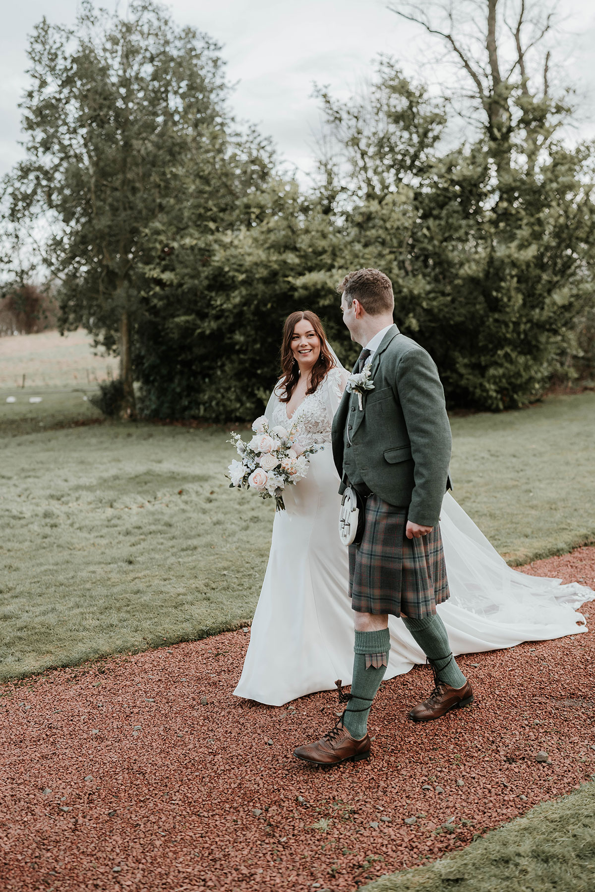 Bride in lace gown and groom in kilt walking hand in hand along red gravel path