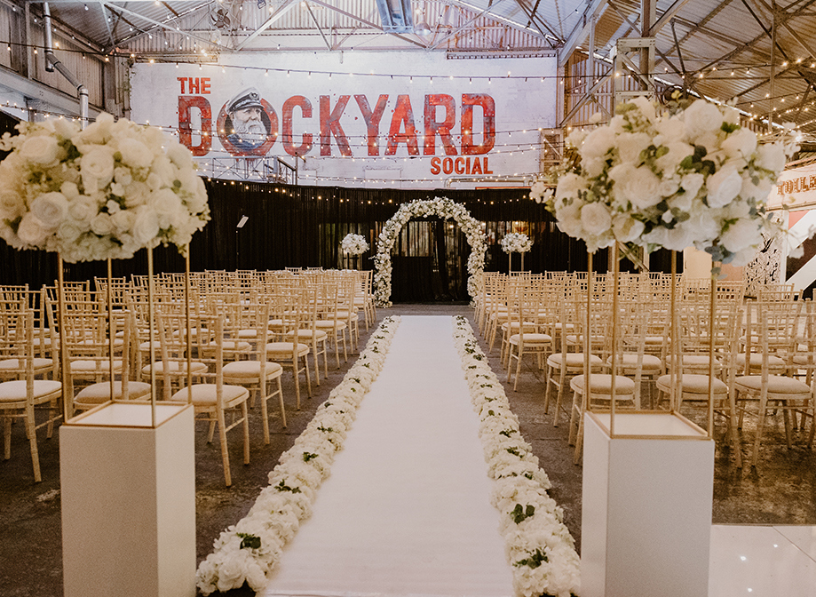 interior of Dockyard Social set for a wedding ceremony with rows of chiavari chairs, white flowers on floor either side of aisle, white pillar flower arrangements and large white floral arch at bottom of aisle. The ceiling is criss-crossed with festoon lights
