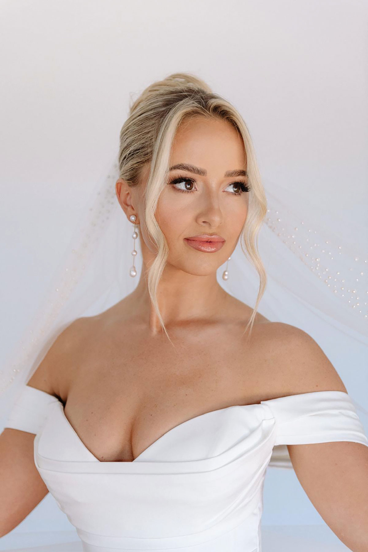 Blonde bride wearing an off-the-shoulder wedding dress and veil, with soft, radiant makeup and glossy skin