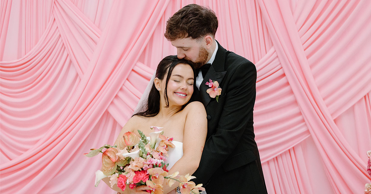 Bride and groom embracing in front of a pink draped backdrop with a vibrant pink and peach bridal bouquet during a wedding at The Collector's Hall in Glasgow