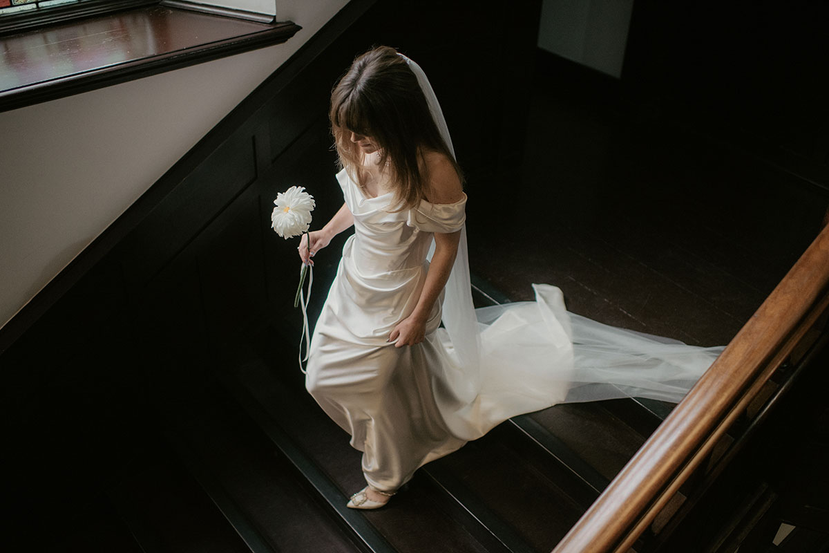 a bride wearing a Vivienne Westwood dress carrying a single stem flower walking down a dark wooden staircase at Achnagairn Castle
