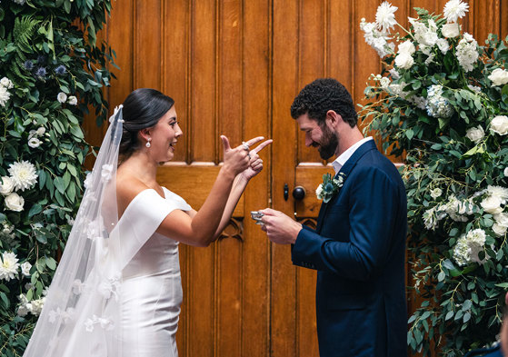 A bride makes a pointed finger gesture while a groom wearing a dark blue suit looks down at a silver quaich vessel in his hands, while standing against a large wooden door at Blairquhan Castle