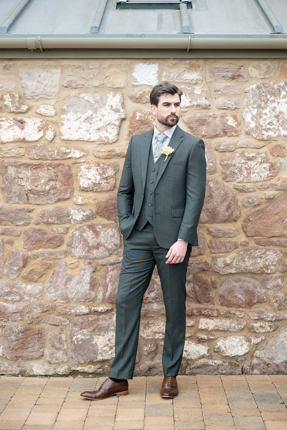 a man wearing a dark-green suit from Slater Menswear standing next to a stone building
