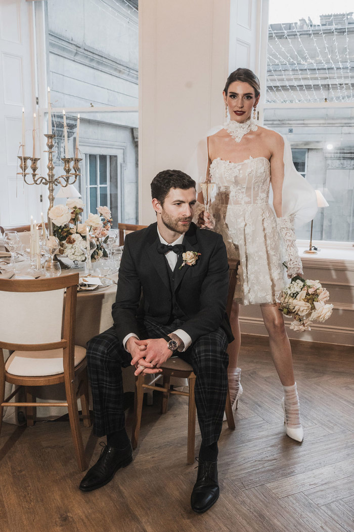 a bride wearing a short lace wedding dress from Eleganza Sposa standing behind a groom wearing a tartan trews outfit from McCalls. They are standing in front of large glass windows and a circular table that has been dressed for a wedding.