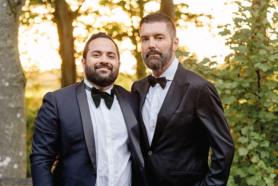 two grooms wearing suits and bowties stand with their arms around each other