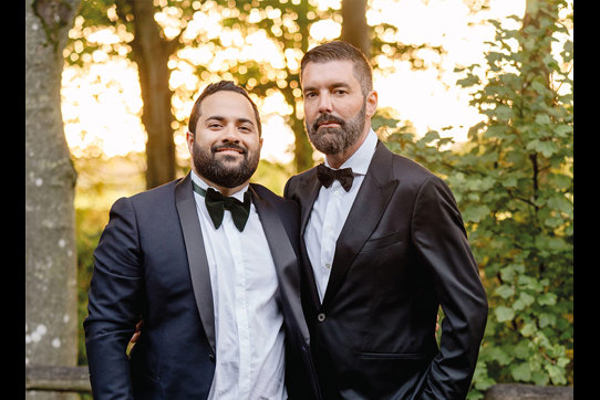 two grooms wearing suits and bowties stand with their arms around each other