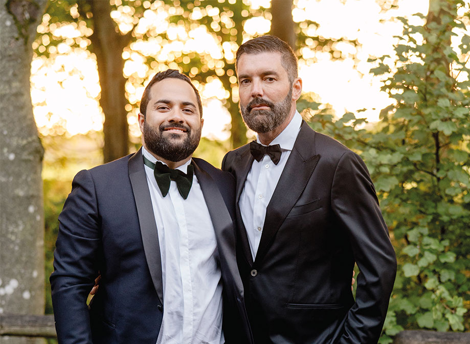two grooms wearing suits and bowties stand with their arms around each other