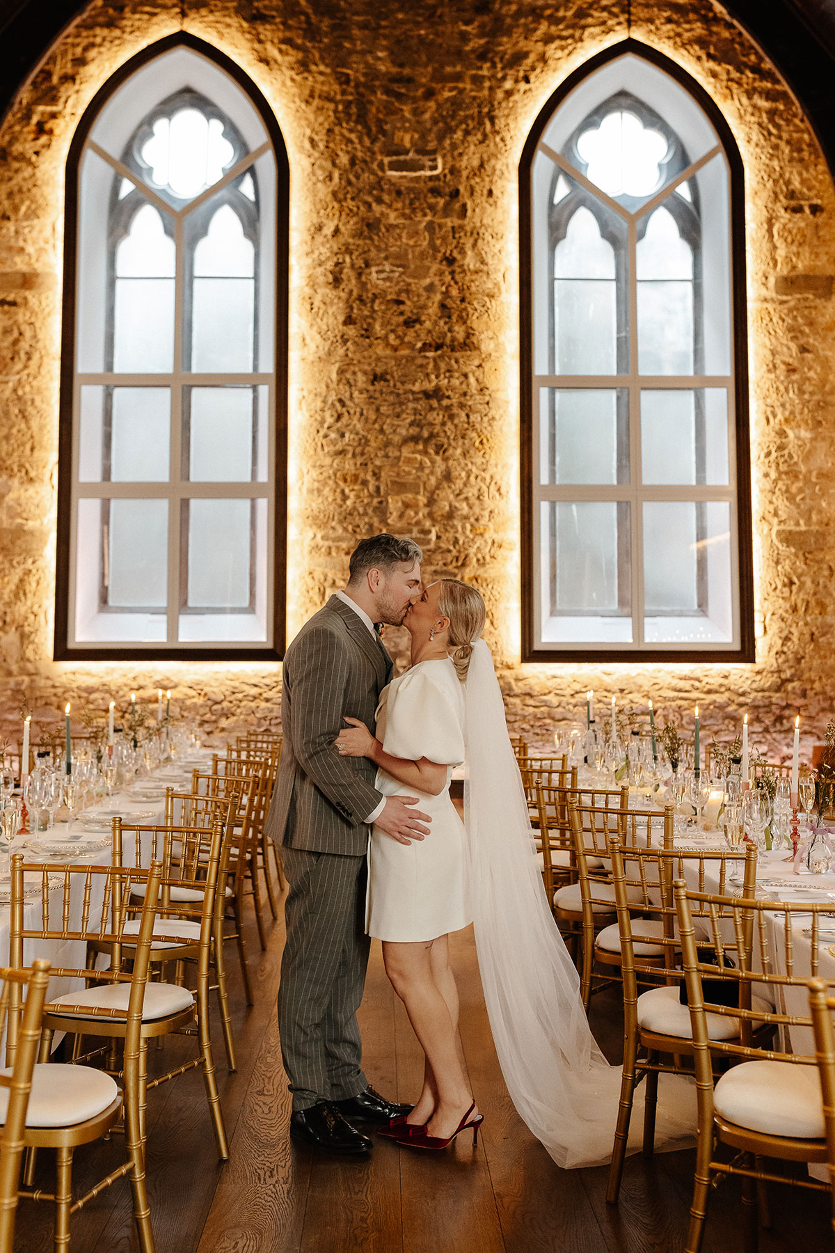 Bride and groom share a kiss between the candlelit dinner tables at Greyfriars Hall, Virgin Hotels Edinburgh, framed by tall arched windows and stone walls.