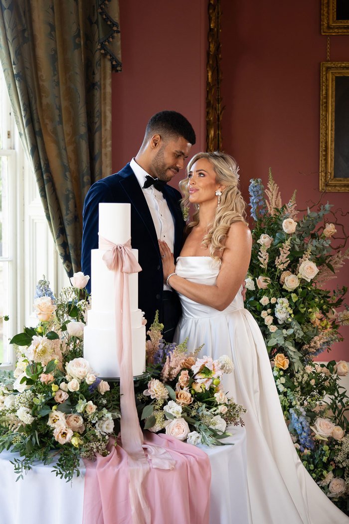 A groom wearing a navy velvet suit jacket looks lovingly at a bride wearing a strapless ballgown wedding dress as they stand in front of a three tier wedding cake with lots of flowers around them