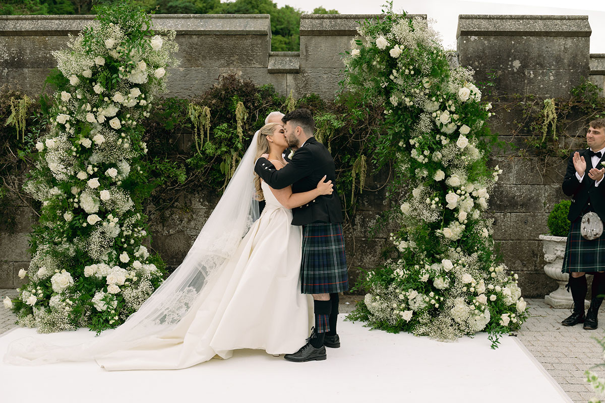 Bride and groom kiss at outdoor ceremony at Dundas Castle with tall white floral pillars and castle stone backdrop.