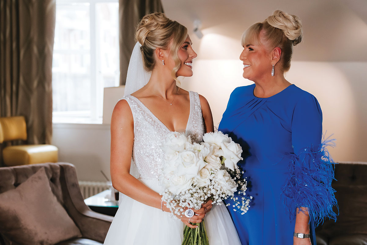 Bride Nicole stands indoors with her mum Carole before the ceremony, both smiling at each other; Nicole wears a sleeveless beaded wedding gown and veil, while Carole wears a royal blue mother-of-the-bride dress with feathered sleeve detail