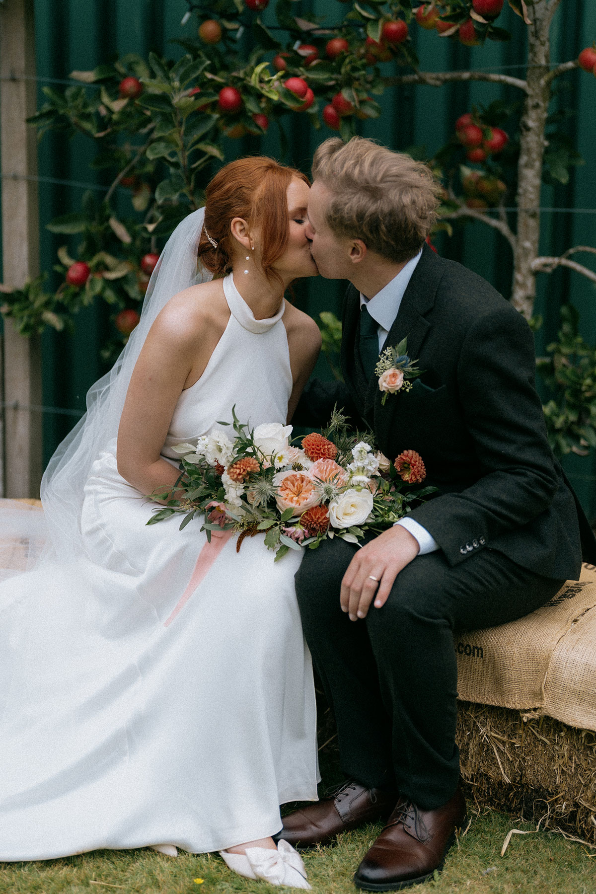 Bride and groom kissing while seated on hay bale surrounded by apple trees and florals