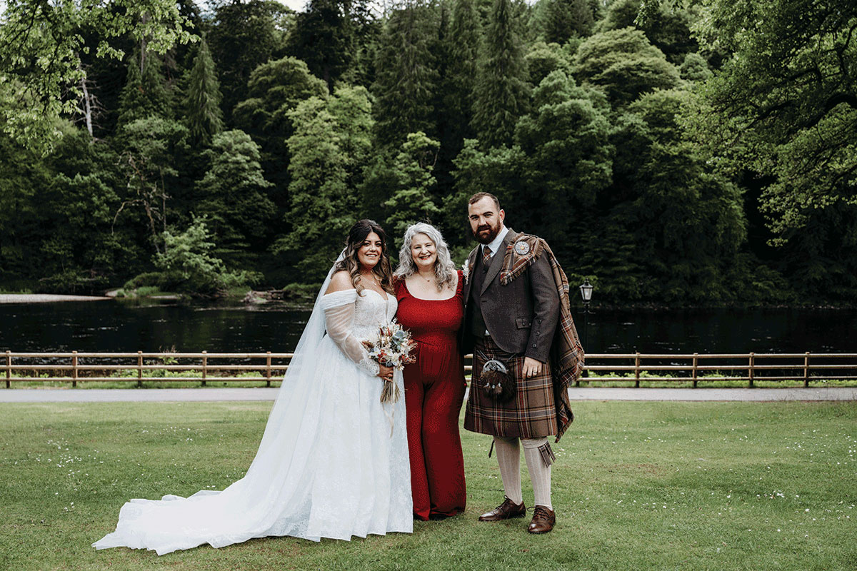 Bride and groom at Dunkeld House Hotel posing with Fuze Ceremonies celebrant Yvonne beside the River Tay and lush green woodland backdrop
