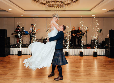 a groom lifting a bride up on a wooden floor. There is a band on a small stage behind them with pyrotechnics.