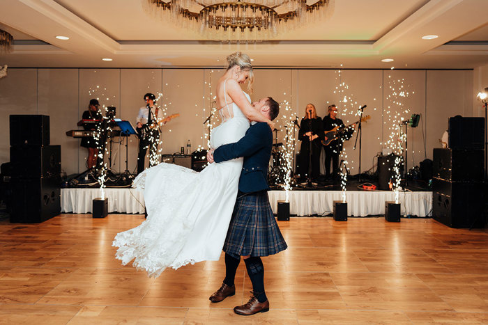 a groom lifting a bride up on a wooden floor. There is a band on a small stage behind them with pyrotechnics.