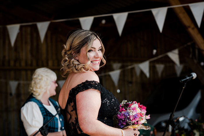 A Smiling Bride Wearing A Black Off Shoulder Wedding Dress Holding A Small Pink And Purple Posy