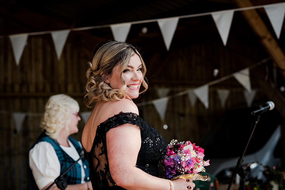 A Smiling Bride Wearing A Black Off Shoulder Wedding Dress Holding A Small Pink And Purple Posy