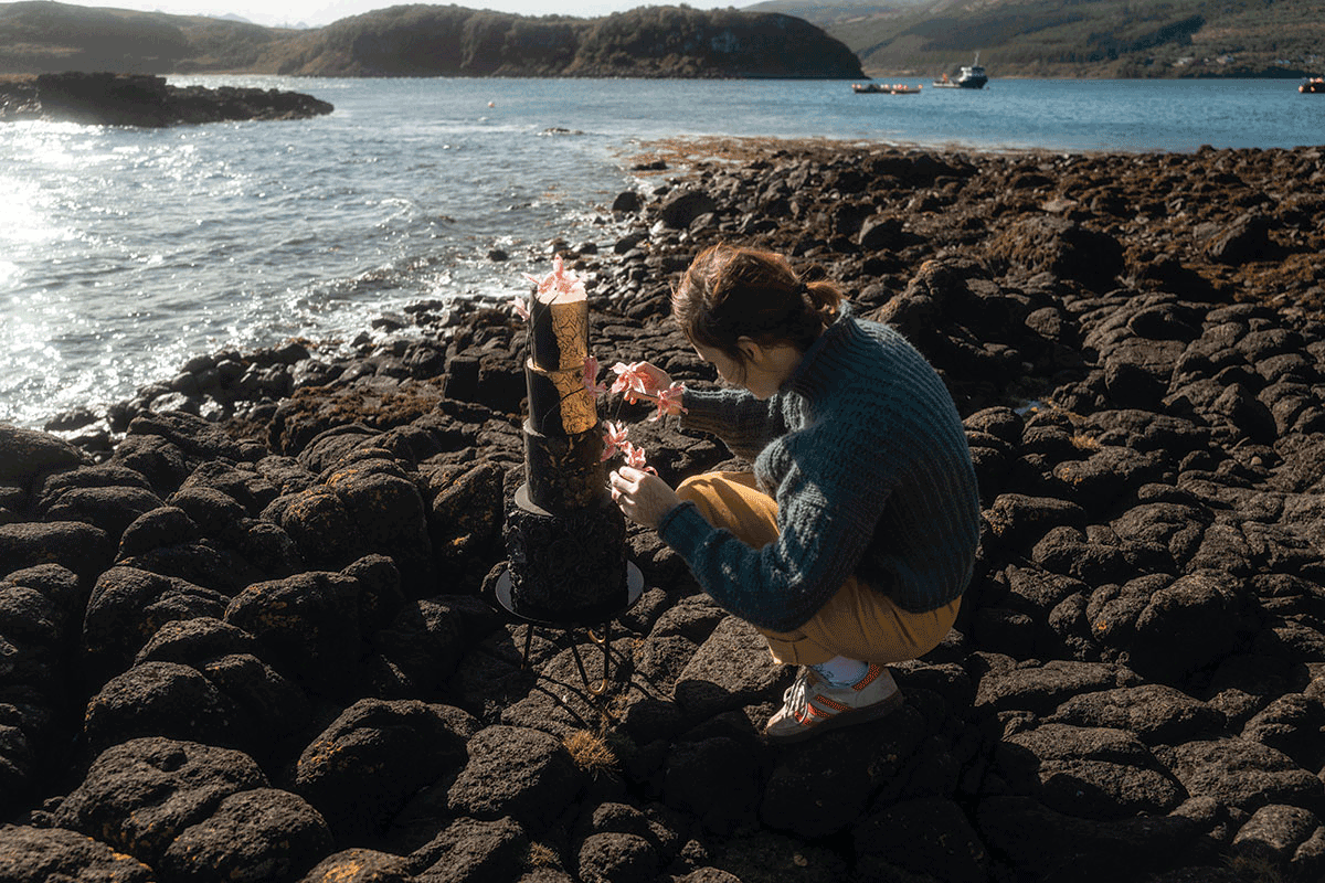 Cake decorator with gold leaf, floral cake in an outdoor seaside setting