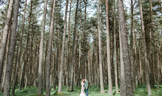 Couple in wedding outfits standing amongst tall trees in forest setting