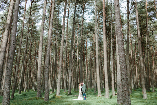 Couple in wedding outfits standing amongst tall trees in forest setting