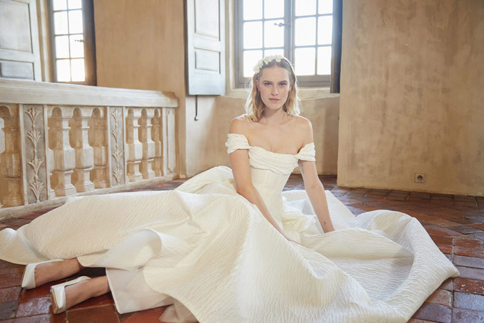 Blonde woman sat on tiled floor wearing a large off the shoulder wedding gown and matching floral headband