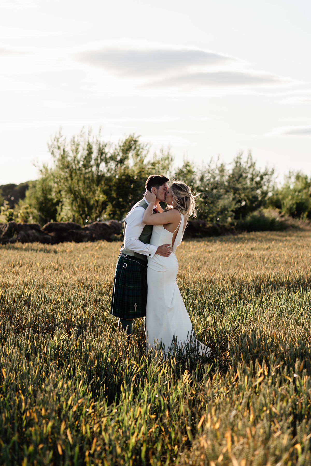 Bride and groom stand close together, kissing in a golden wheat field at sunset, with soft trees and sky behind them