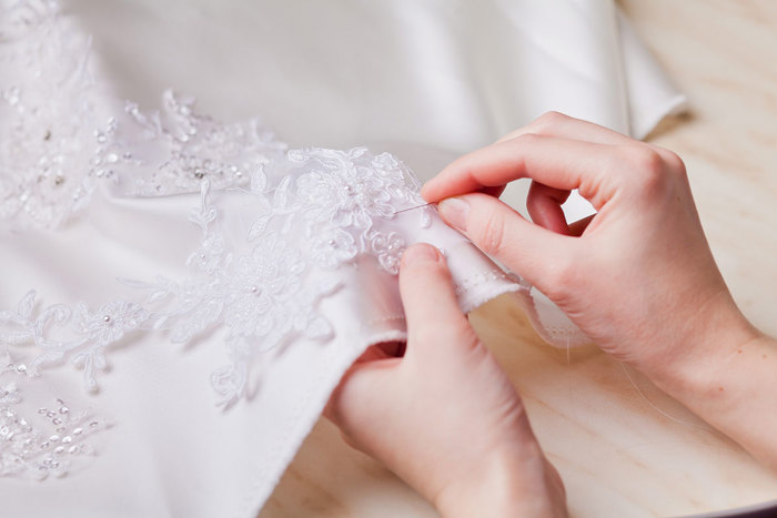 an up close view of a pair of hands holding a sewing needle along the hem of a white dress embroidered with floral decorations 