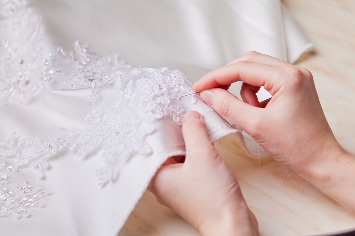 an up close view of a pair of hands holding a sewing needle along the hem of a white dress embroidered with floral decorations