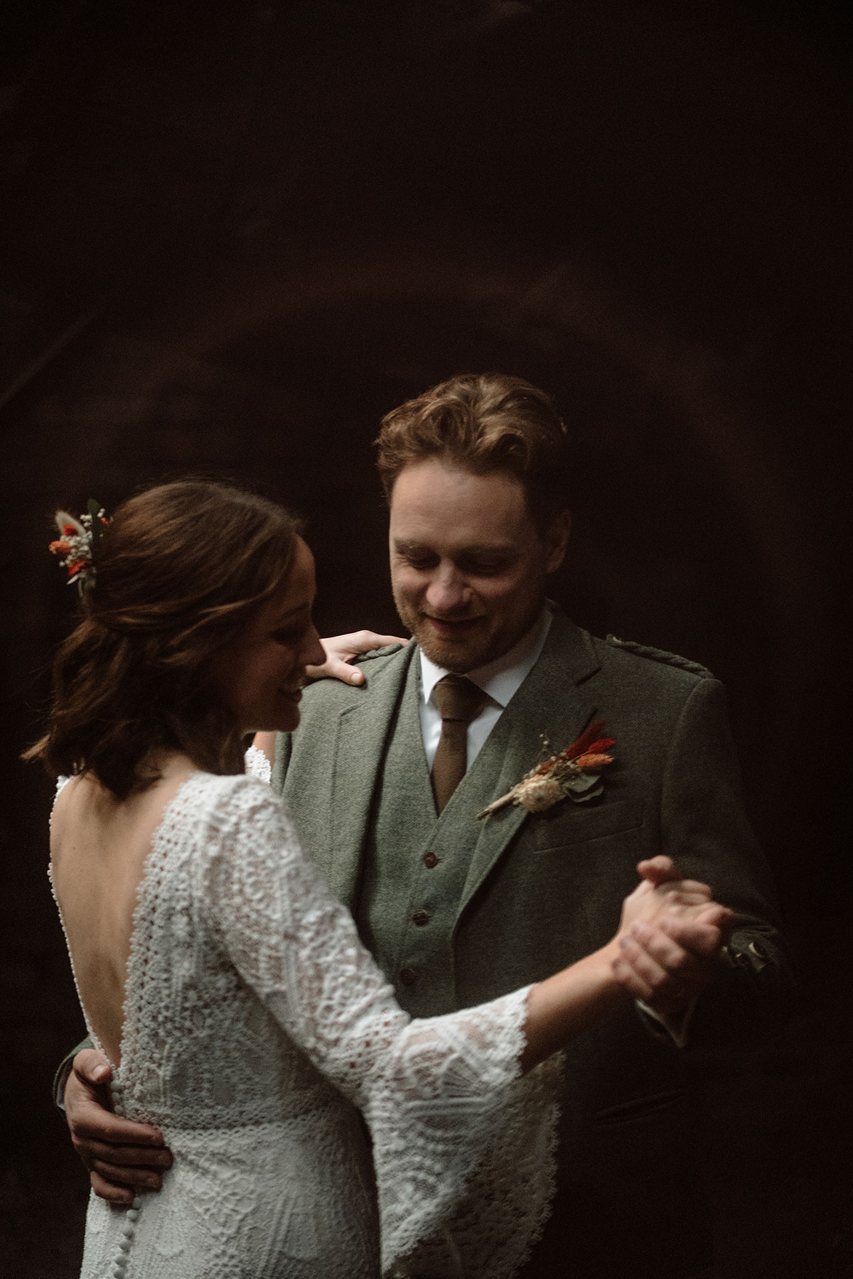 Bride and groom share their first dance beneath warm lighting inside an industrial wedding venue, with the bride wearing a long-sleeved lace wedding dress and the groom in a tweed kilt outfit