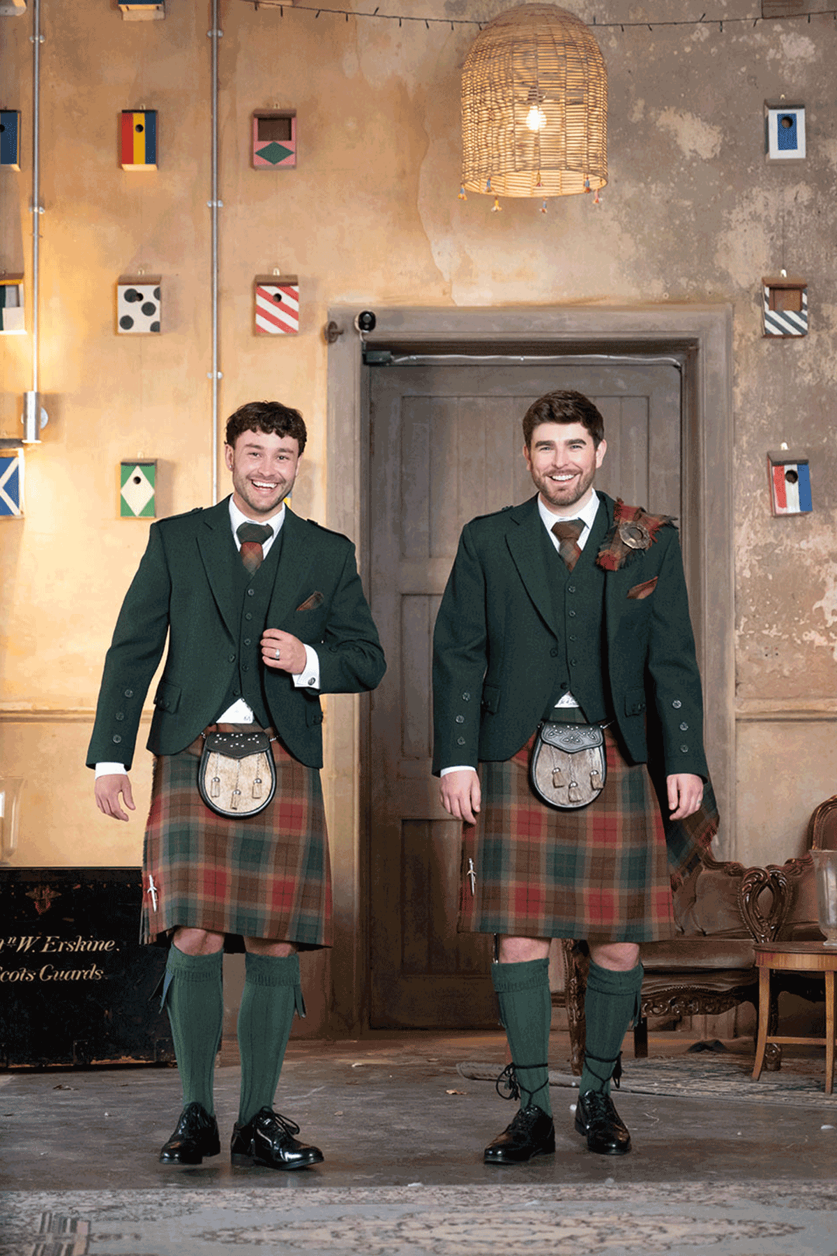 Two grooms wearing contemporary tartan kilts and dark green Highlandwear walking through stylish rustic interiors during a Scottish groomswear editorial shoot